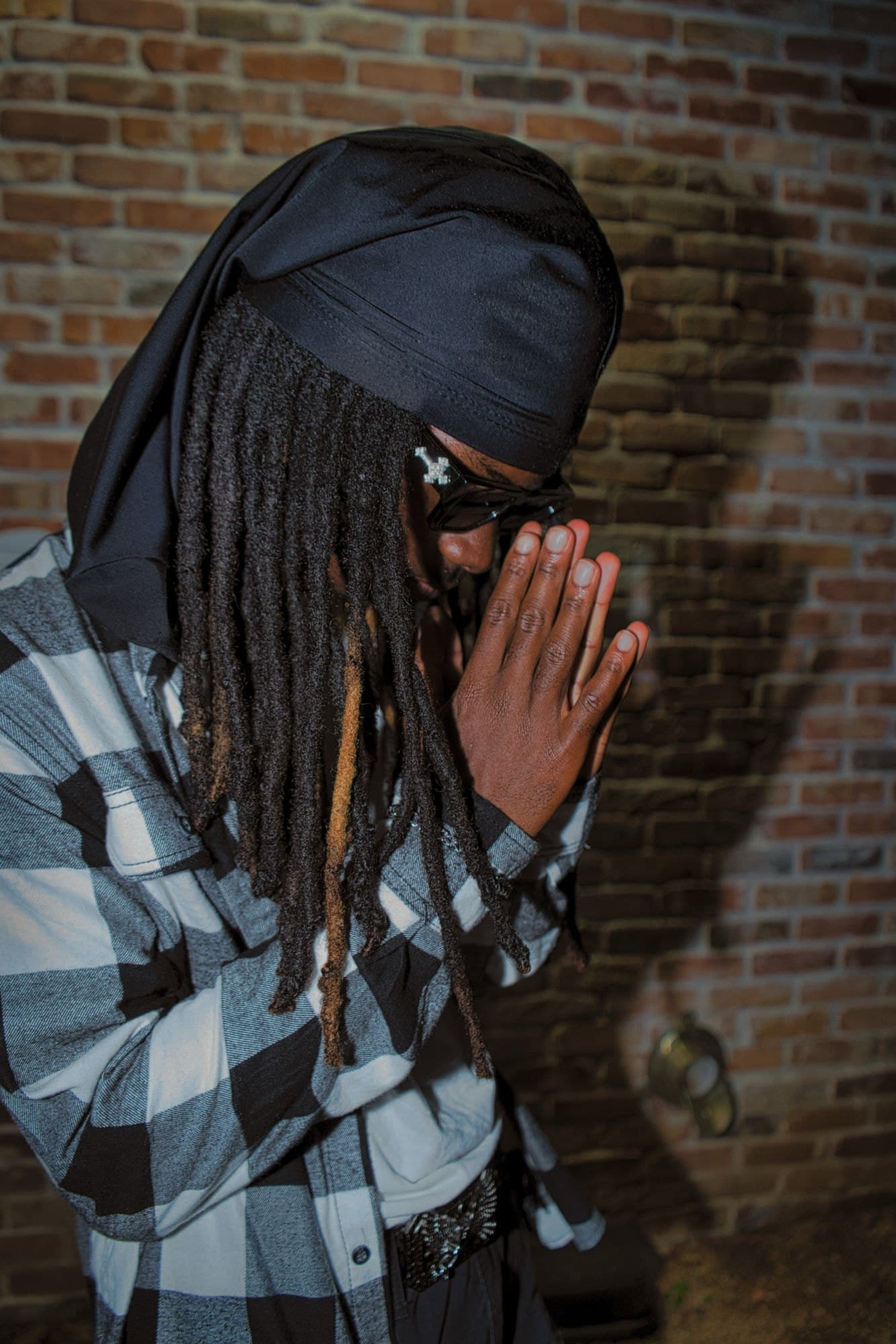 Direct flash portrait against a brick wall, hands together, locs and durag