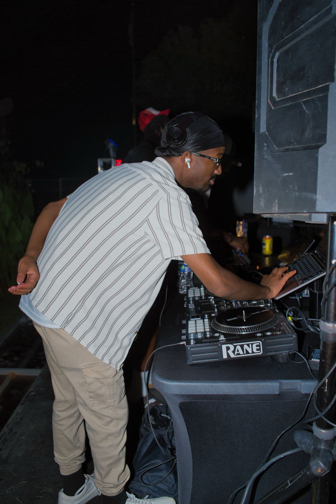 DJ leaning into the decks beside a stack of speakers at an outdoor set
